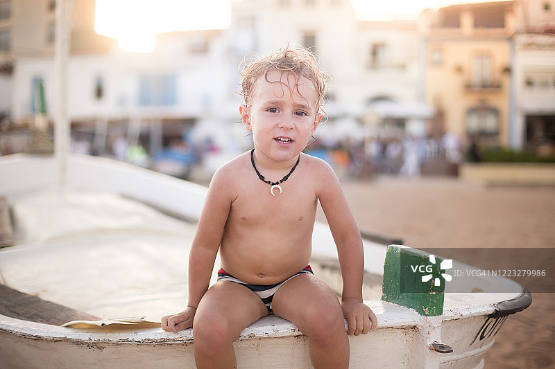 可爱的男孩坐在沙滩上的船上看镜头图片素材
