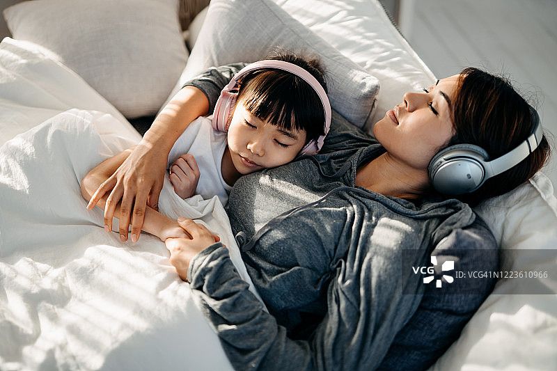 放松的年轻亚洲母亲和小女儿闭着眼睛躺在床上，用耳机听音乐图片素材