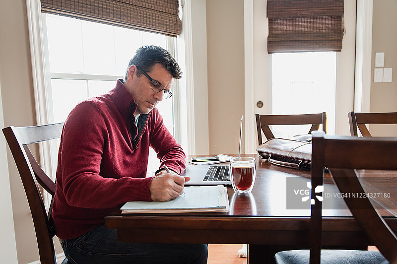 戴眼镜的男人在家工作，在餐桌上使用电脑。图片素材