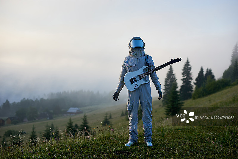 清晨，穿着太空服的宇航员拿着吉他站在美丽的青山草地上图片素材