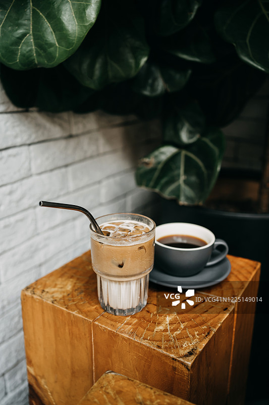一杯冰拿铁和一杯咖啡放在咖啡店的木桌上，靠着白砖墙和盆栽图片素材