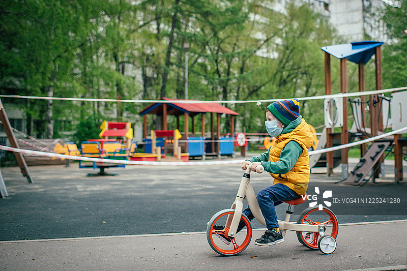在因新冠肺炎疫情而关闭的游乐场附近，戴着防护面具的小男孩骑着跑步自行车图片素材