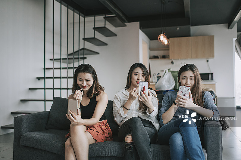 3名亚洲华人女性在客厅沙发上用手机当镜子涂口红图片素材