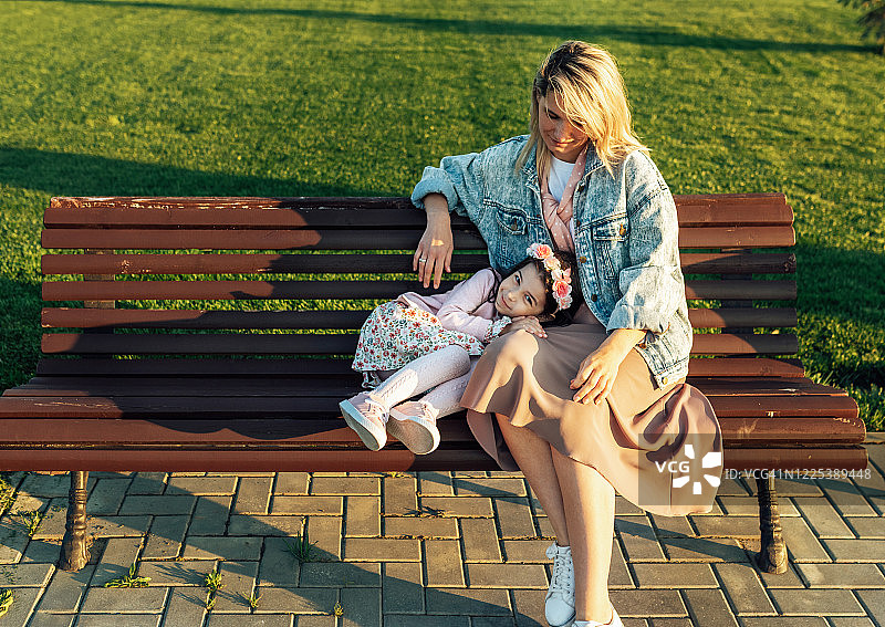 全身像美丽的妈妈坐在长椅上和她可爱的小女孩在公园。美丽的年轻女人放松和她的孩子，享受在一起的时间。母亲和女儿分享爱图片素材