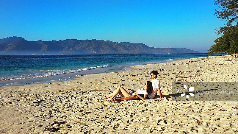 一对浪漫的情侣坐在巴厘岛的白色沙滩上，放松地看着清晨的日出。图片素材