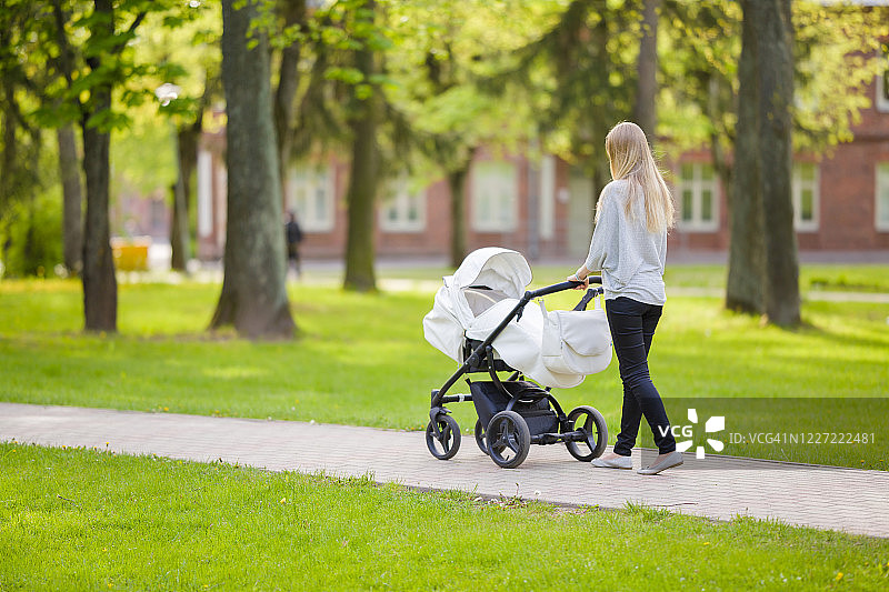 一位年轻的母亲推着一辆白色婴儿车，在温暖、阳光明媚的春天里，慢慢地走在镇绿公园里。花时间陪婴儿，呼吸新鲜空气。享受漫步。图片素材