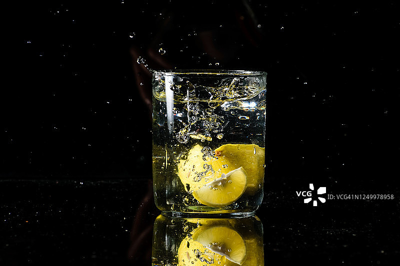 将一杯水放在有黑色背景的反光表面上，将一个柠檬倒入杯中，水就会飞溅。夏天的概念图片素材