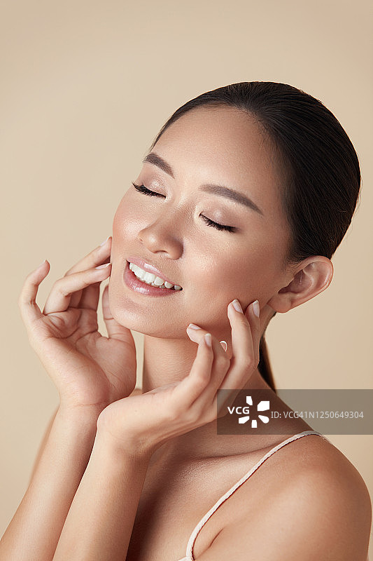 皮肤护理。亚洲模特肖像。年轻女子闭着眼睛触摸脸部米色背景。女性享受健康的皮肤在日常美容和美容治疗后。图片素材