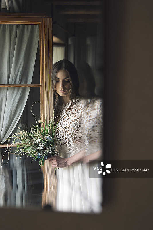 年轻女子在优雅的婚纱拿着花束看着窗户图片素材