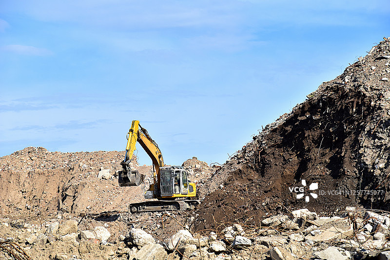 黄色挖掘机在堆填区弃置建筑废物。在蓝天的背景下，挖土机在采石场挖掘砾石。回收拆除后的混凝土和沥青。图片素材