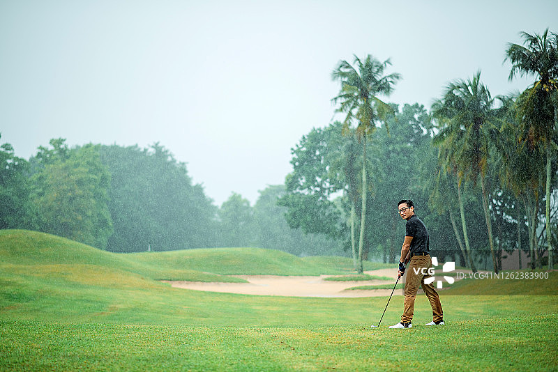 在雨天的高尔夫球场上，亚洲中国年轻男性高尔夫球手准备开球，挥动他的高尔夫球杆图片素材