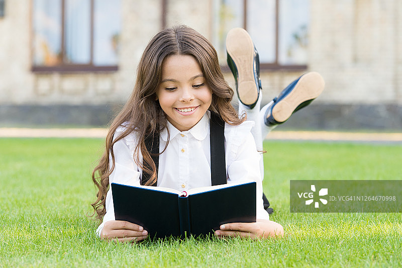 基础教育。可爱的小女孩学习阅读。穿着校服的女生躺在草坪上，旁边放着最喜欢的书。学习的概念。课外阅读。可爱的小孩在户外看书图片素材
