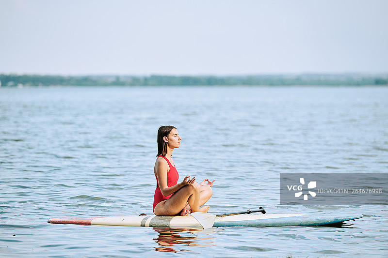 年轻的女运动员在红色泳装练习瑜伽，坐在冲浪板上图片素材