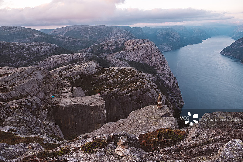 阴天里的挪威峡湾，蓝色的露营帐篷和身穿红色夹克站在悬崖上的不可辨认的人。图片素材