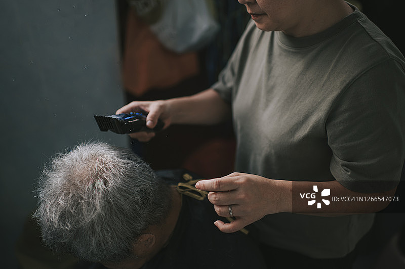 由于旅行禁令，一位亚洲华人中年妇女正在厨房里为她的父亲修剪头发图片素材