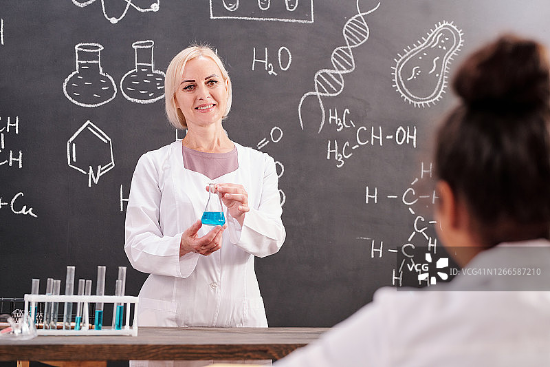 年轻的金发微笑的化学老师拿着一个装有蓝色液体物质的试管图片素材