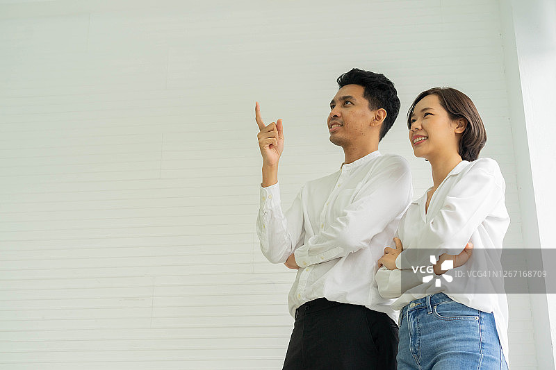 近距离的亚洲女子手臂交叉，站在男人旁边，指着手在白色的墙壁背景内的新房子为恋人夫妇的住房贷款和房地产投资的概念图片素材