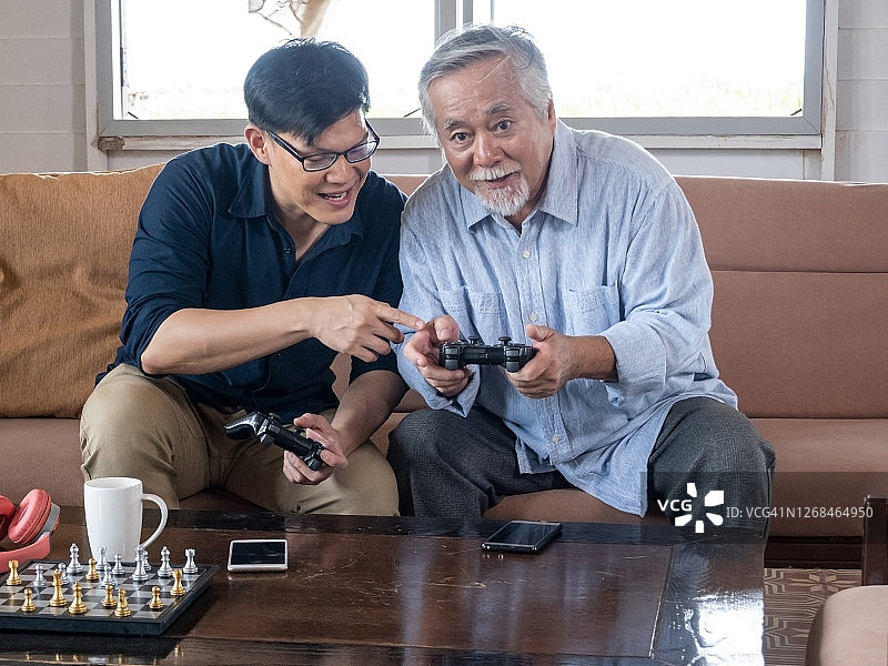 年长的亚洲男人和他的儿子在客厅玩掌机游戏，呆在家里，幸福的微笑。图片素材