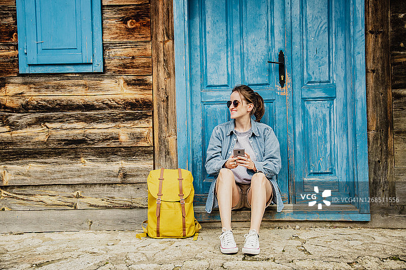 一个背着背包的女人坐在蓝色的门前，用着智能手机图片素材