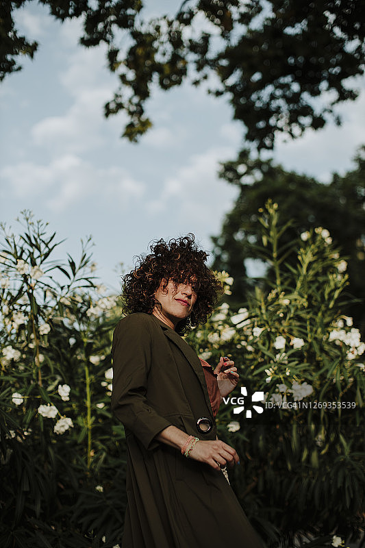 一头卷发的中年妇女，穿着外套，靠着公园里的植物站着图片素材