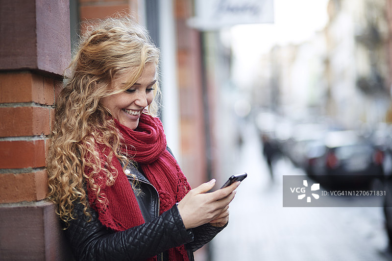 微笑的年轻女子使用手机靠在一堵砖墙在城市图片素材