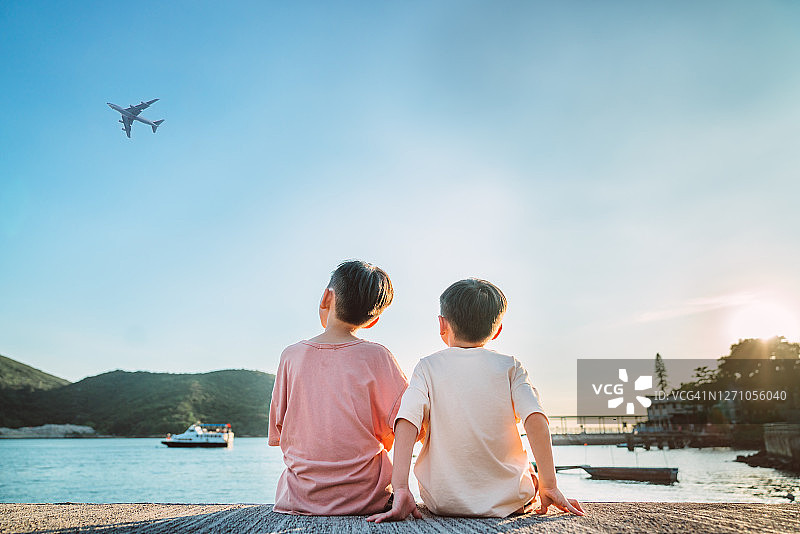 两个小兄弟肩并肩坐在码头上看日落时的飞机。图片素材