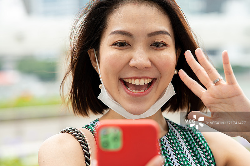 东南亚。新型冠状病毒肺炎技术。网络。美丽微笑的亚洲妇女的肖像与保护面具看智能手机，与朋友视频通话图片素材