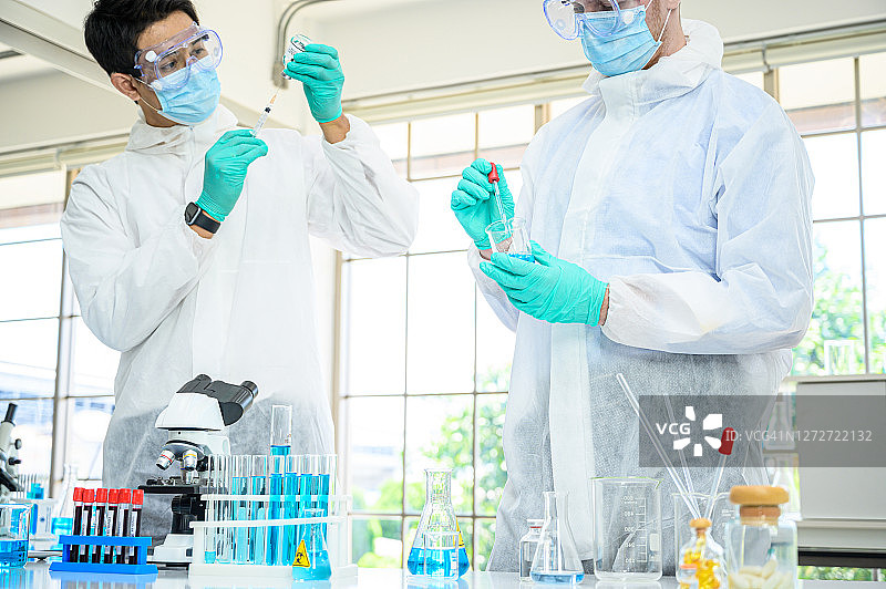 一对男性科学家穿着防护服，手持疫苗，在实验室使用许多实验室设备研究疫苗。图片素材