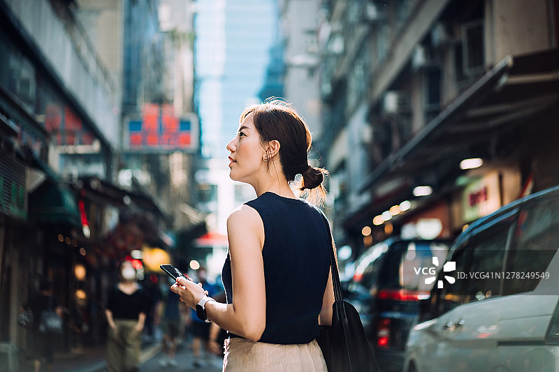 美丽的年轻亚洲女子仰望天空，手里拿着智能手机走在城市街道上，在城市中访问和探索图片素材