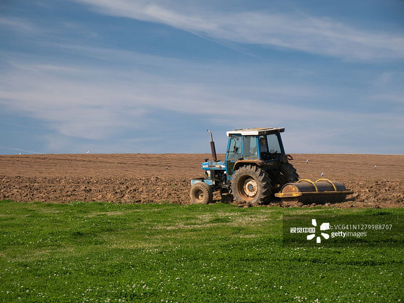 在一个平静、阳光明媚的夏末，一个农民在拖拉机上犁地，准备播种作物。图片素材