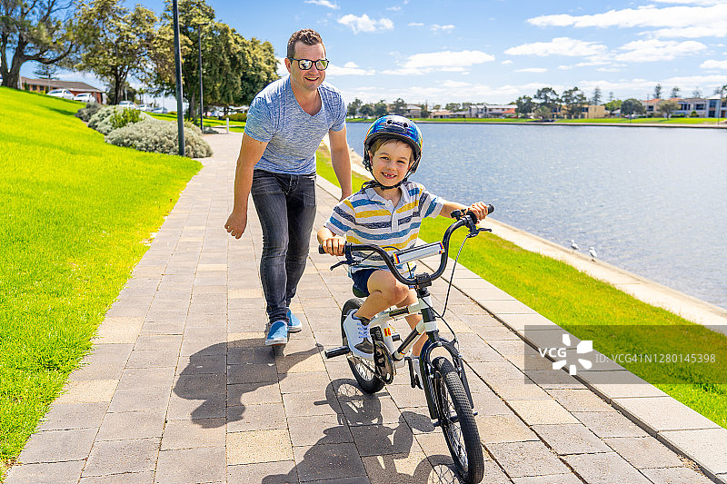 在湖边的公园里，男孩和他的父亲一起学骑自行车。父亲和儿子在自行车上一起玩。幸福家庭，户外活动，童年和育儿理念。图片素材