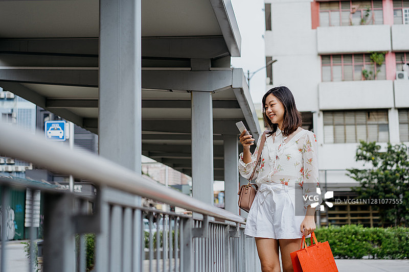一名拿着购物袋使用智能手机的亚洲华裔女性图片素材