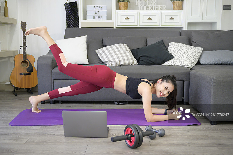 一个女人正在做瑜伽平板支撑，在她的笔记本电脑上看在线培训教程，在家里健身，保健技术概念。图片素材