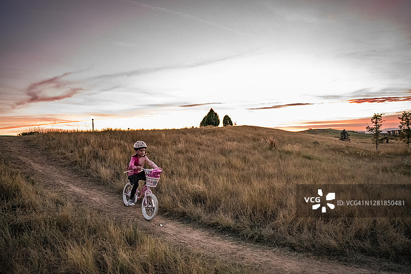 日落时分，年轻的女孩骑着自行车从长满草的小山上下来图片素材