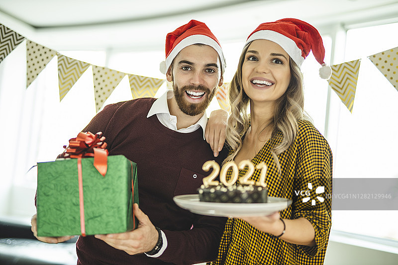 年轻夫妇戴着圣诞帽，拿着圣诞礼物和巧克力蛋糕，在家庆祝新年图片素材