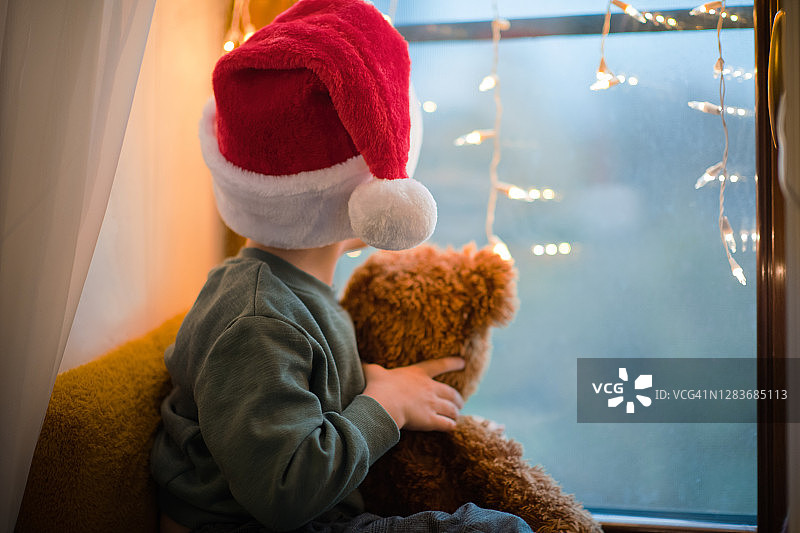 可爱的小男孩在家庭隔离戴着圣诞帽和他的泰迪熊在圣诞节图片素材