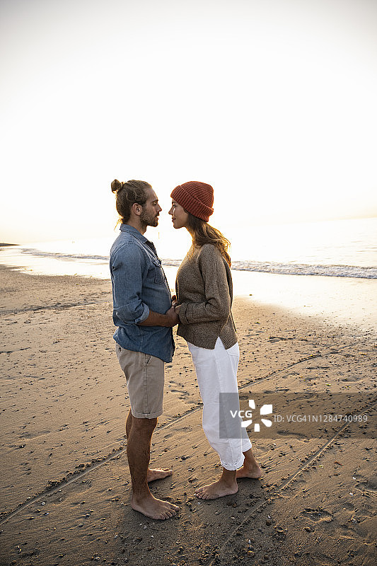 在日落时分，一对浪漫的年轻情侣手牵着手站在岸上，看着对方图片素材