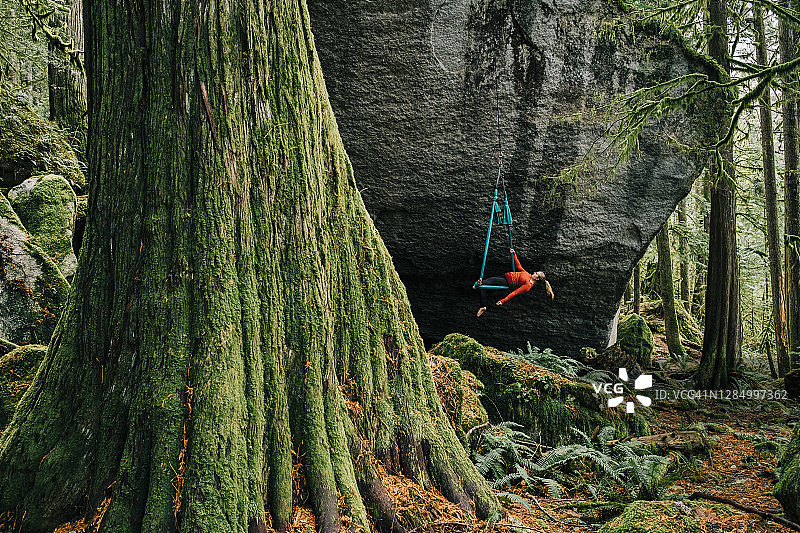 一个年轻的成年女性在一个悬挂在巨大岩石表面的吊床上表演杂技动作图片素材