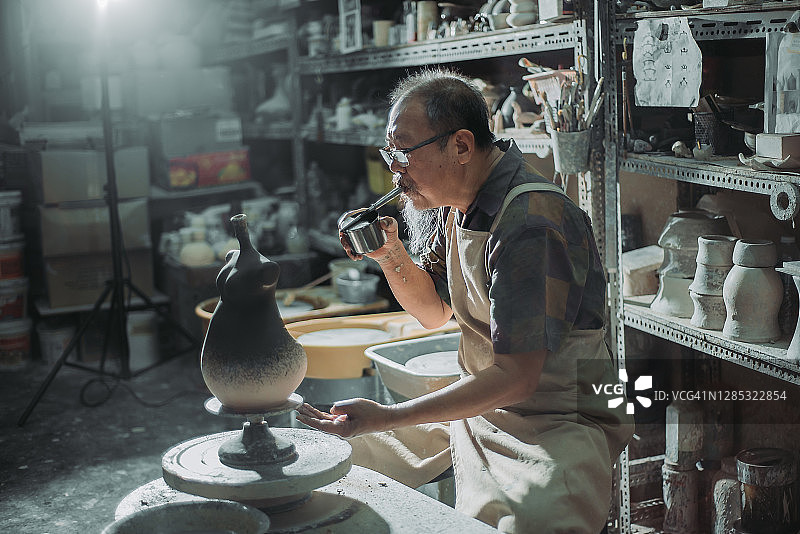 亚洲华人高级陶艺师在他的工艺工作室里在陶器上吹颜料图片素材