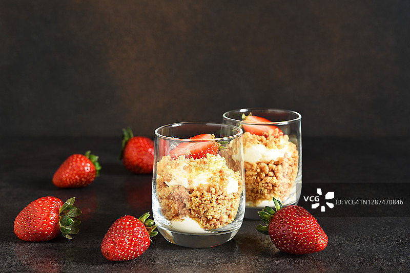 奶酪蛋糕装在玻璃杯里，草莓放在深色的混凝土背景上。杯子里的甜点。图片素材