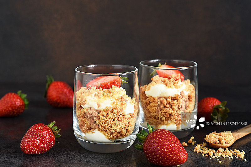 奶酪蛋糕装在玻璃杯里，草莓放在深色的混凝土背景上。杯子里的甜点。图片素材