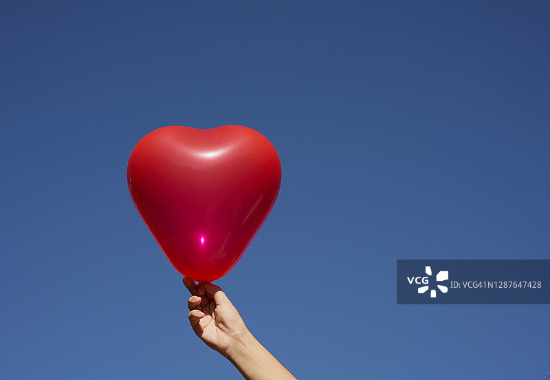 a white hands holding a red balloon with heart shape as a conceptual idea of a strong healthy heart, a transplant heart or saint valentine´s day with blue sky background. Horizontal photo.图片素材