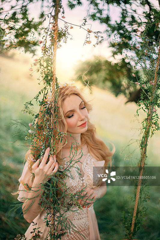 年轻快乐的金发女子形象的森林仙女在一个浅桃复古连衣裙，站在一棵树附近。时尚模特女孩摆姿势在夏季绿色的自然背景，日落的太阳。长长的金发。图片素材