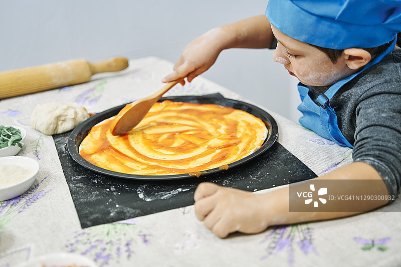 一个男孩打扮成厨师和他的母亲在披萨上添加番茄酱的特写镜头图片素材