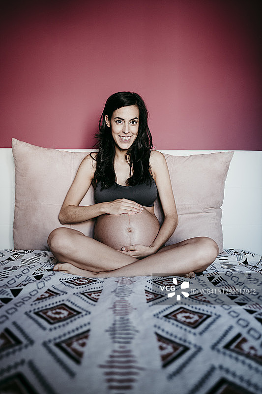 面带微笑的孕妇坐在家里靠红墙的床上图片素材