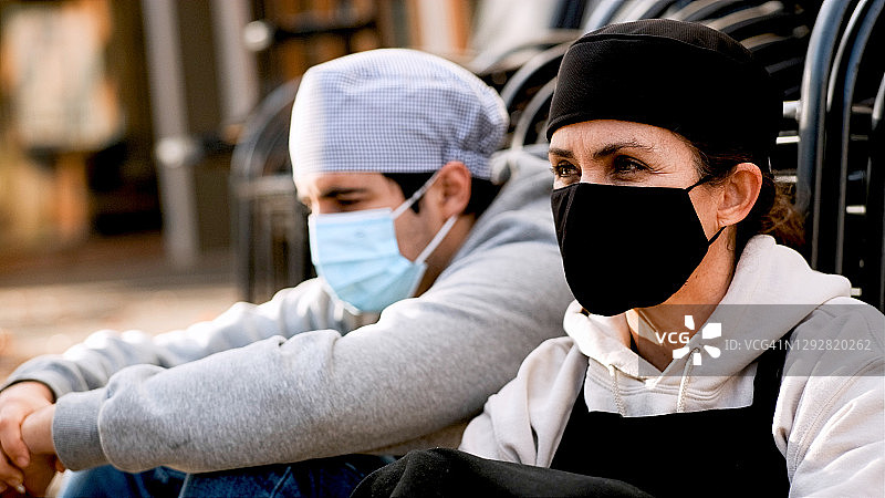 一名成熟的女厨师和一名戴着防护口罩的年轻男同事坐在他们工作的餐厅外面的地板上图片素材