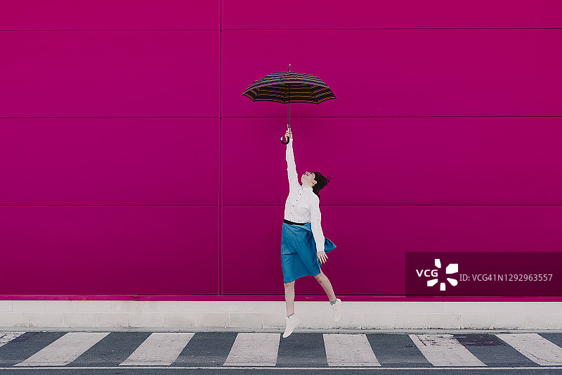 年轻女子拿着伞在一堵粉红色的墙前跳着图片素材