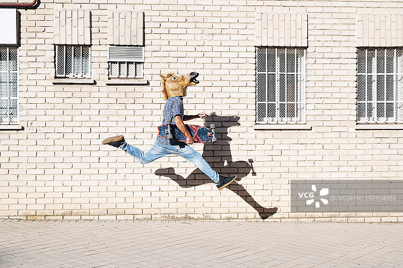 在阳光明媚的日子里，一名男子戴着马面具，手持滑板在城市建筑的人行道上跳跃图片素材