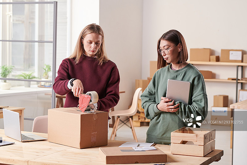 严肃的年轻女仓库员工穿着毛衣站在办公室的木桌旁，用胶带包装包装图片素材