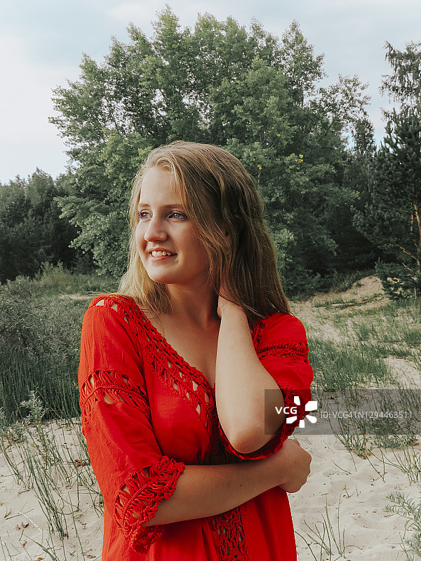在一个阳光明媚的夏天，一个金发的年轻女子在绿色的森林边上快乐地笑着，她穿着红色的裙子图片素材